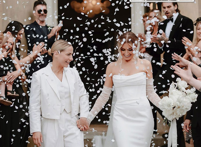 two brides holding hands as white confetti is thrown over them by guests in black standing on either side of them