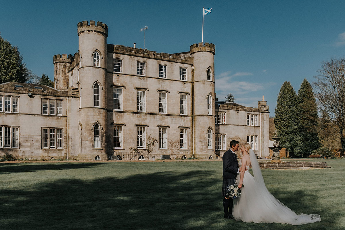 Newlyweds embracing on the grounds of Melville Castle, with the historic Scottish castle and fountain creating a backdrop for wedding portraits