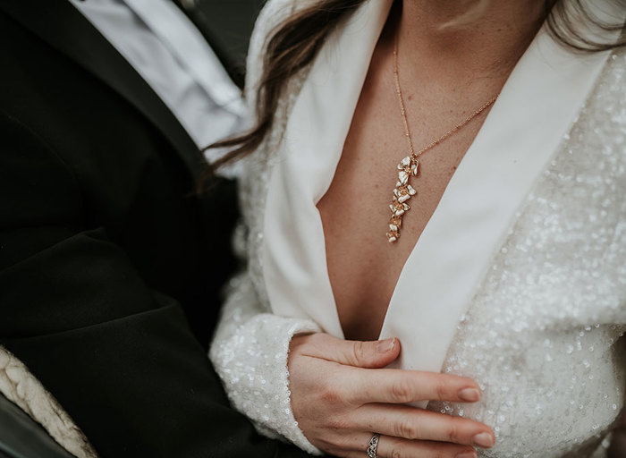 A close up of a couple standing next to each other, the man in a black suit and the woman in a white sparkly suite with a long pendant necklace
