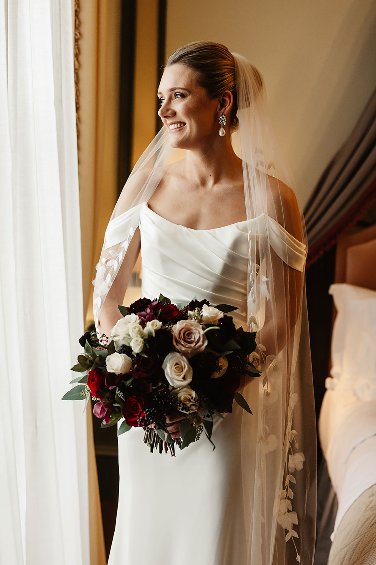 Bride smiling while standing beside a window, holding a bouquet of red, blush and burgundy flowers.