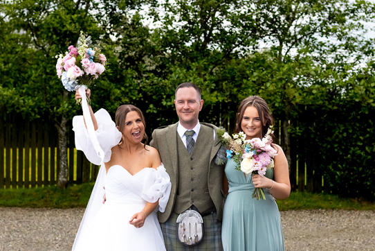 bride standing in white dress holding flowers above head, groom in kilt and young girl in teal dress