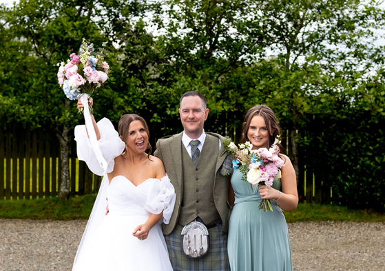 bride standing in white dress holding flowers above head, groom in kilt and young girl in teal dress