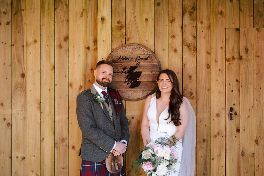 A bride and groom stand in front of a wooden wall and a wooden sign that says 'Helen + Grant' with a map of Scotland