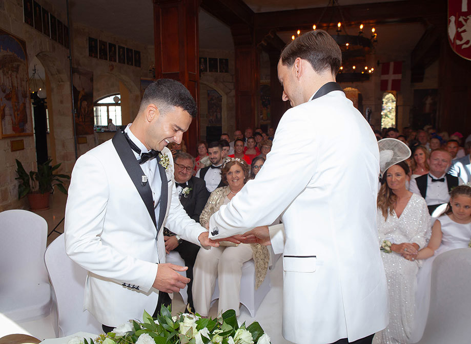 two grooms exchanging rings in a dark stone room as seated wedding guests watch on