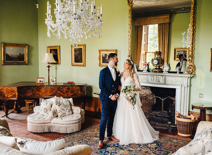 Bride and groom stand in front of unlit fireplace and a wooden piano in a large green room