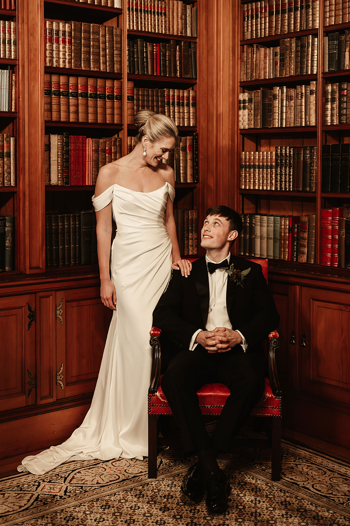 Bride standing beside the groom seated in a red chair, surrounded by tall wooden bookshelves in a historic library.