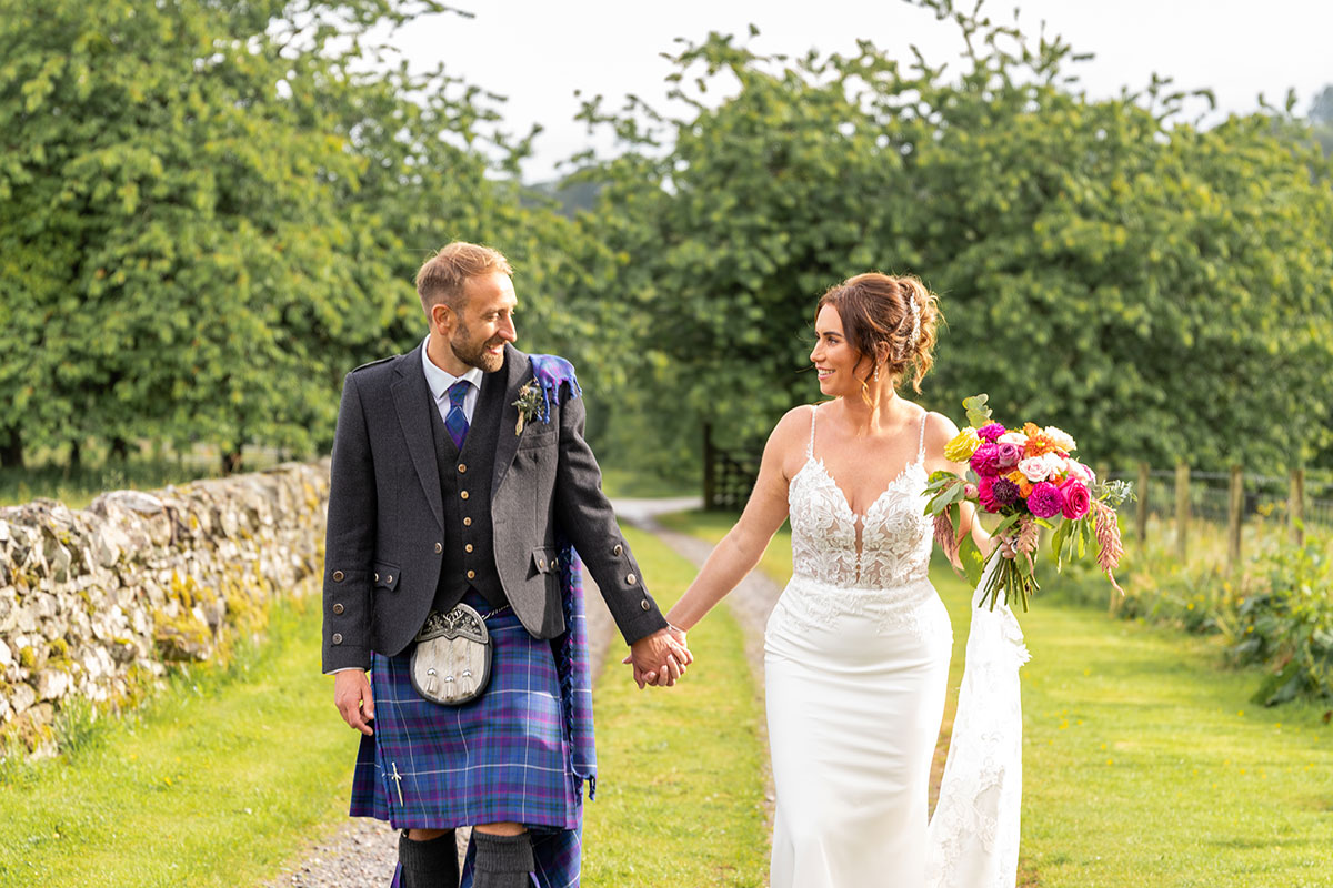 Bride holding a colourful bouquet walks hand in hand with her groom in a tartan kilt beside a stone wall at Ardoch Loch Lomond.