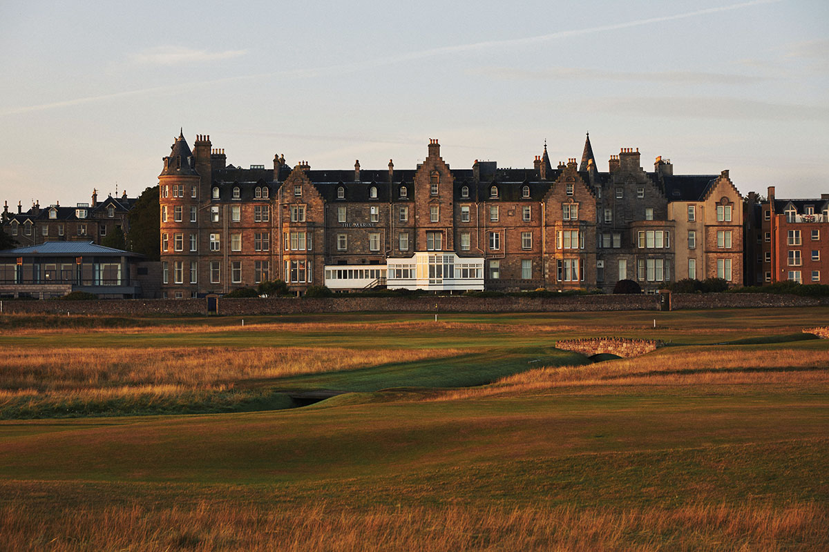 Large stone hotel building viewed across a golf course at golden hour, with warm light on the façade and low grasses in the foreground