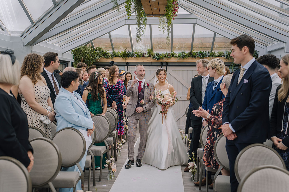 Bride walking down the aisle with her father during an indoor glasshouse wedding ceremony surrounded by guests