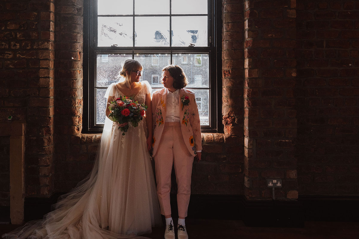 Two brides standing by a large industrial window, holding a colourful bouquet and looking at each other in natural light.
