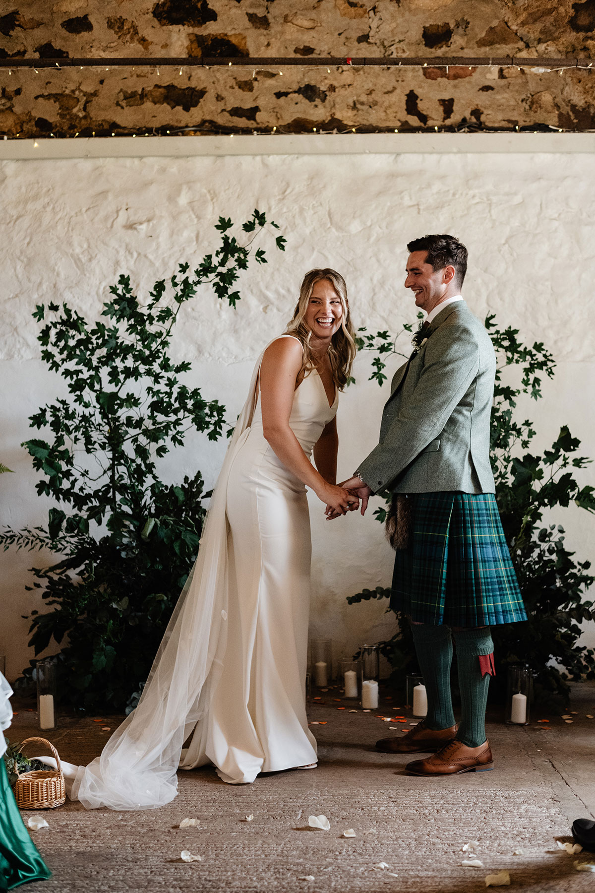 Bride and groom holding hands and laughing during their ceremony against a backdrop of greenery and candles