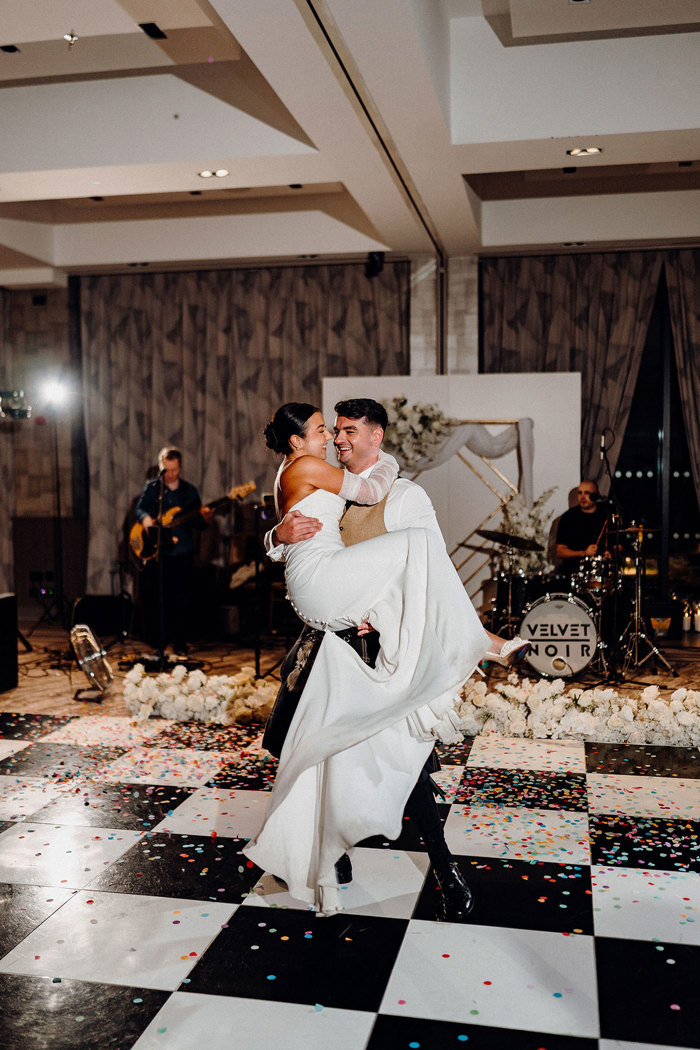 bride in strapless wedding dress with detached tulle sleeves is held in the arms of tall groom in beige and dark green kilt outfit on a tiled and confetti covered dancefloor