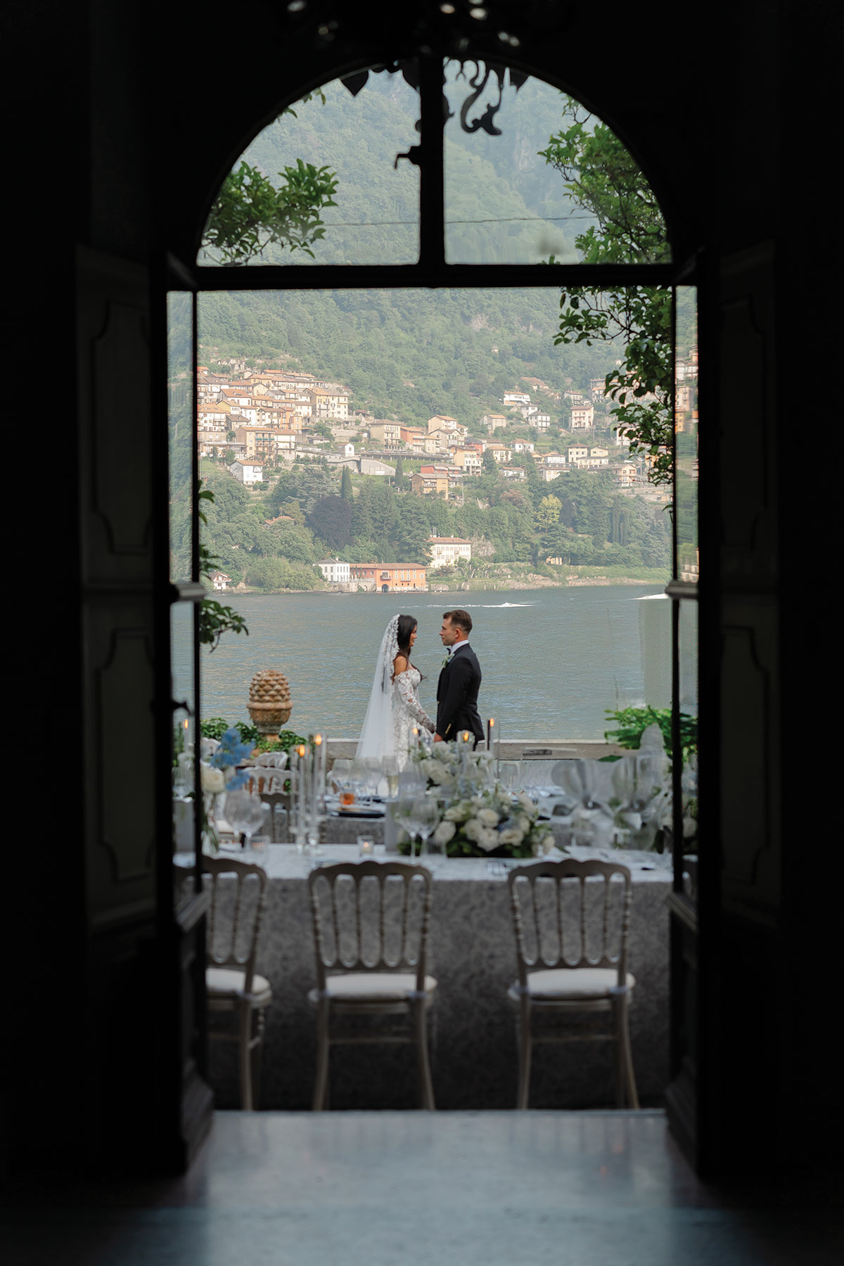 Bride and groom standing by Lake Como, framed through a doorway, with an outdoor reception table set for dinner overlooking the water