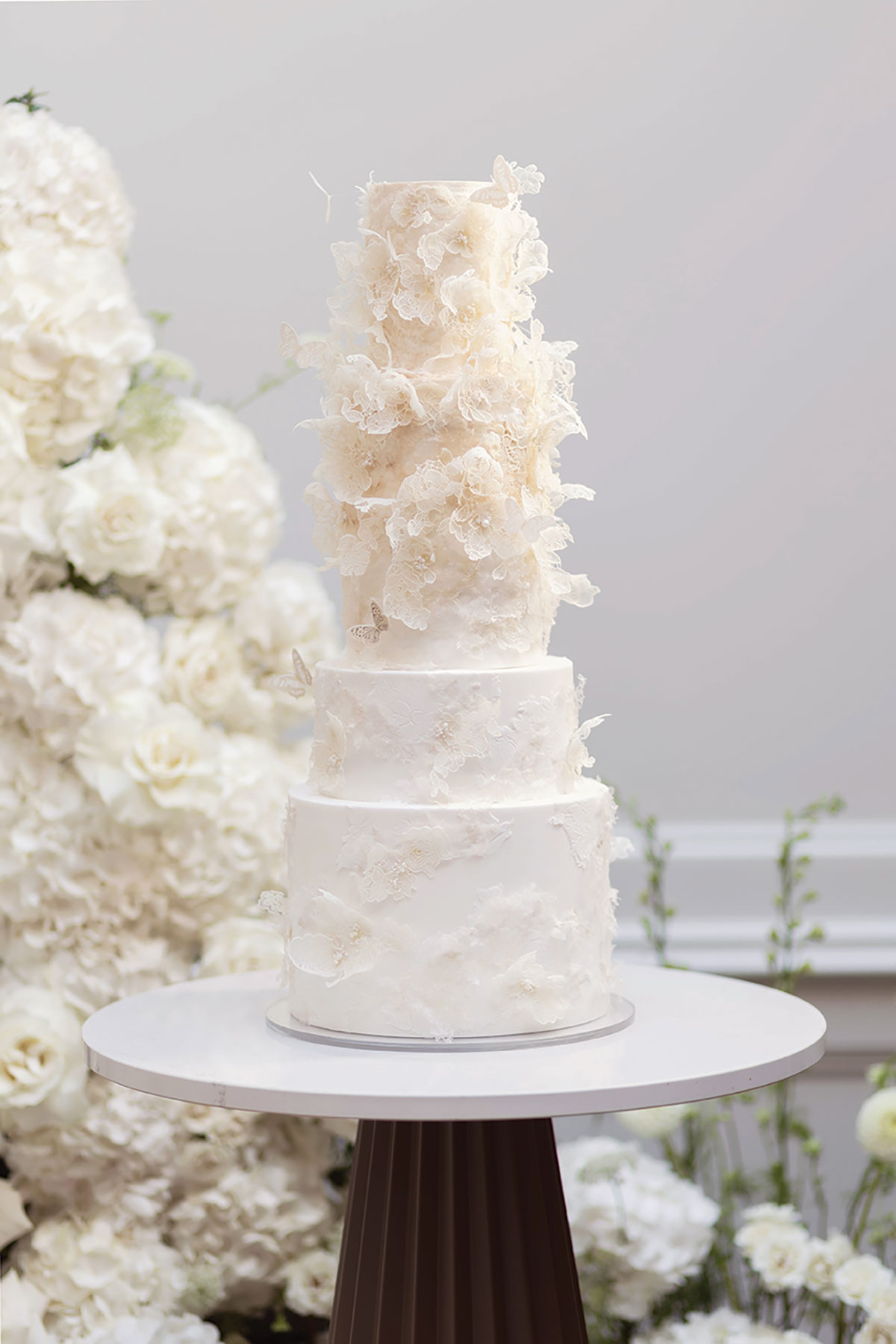 Tall tiered white wedding cake with lace-effect florals and butterflies, surrounded by white flowers