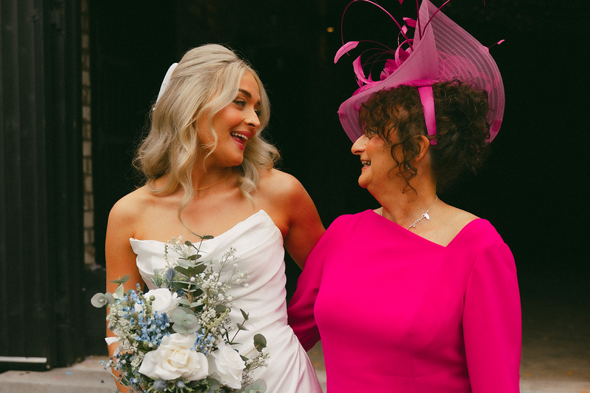 Bride in a strapless white gown holding a bouquet of white and blue flowers smiles at an older woman in a bright pink dress and large fascinator as they stand outside a dark barn doorway