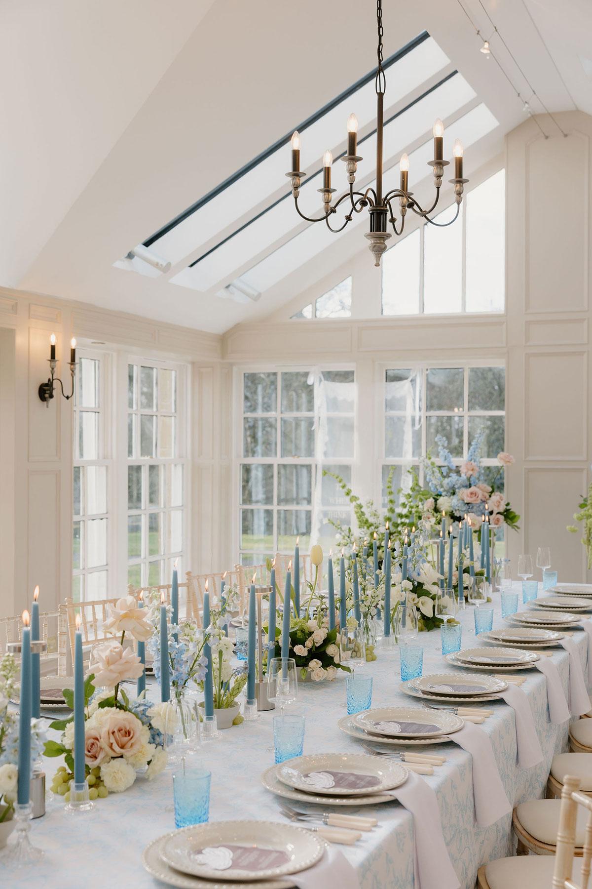 Long reception table with blue candles, pastel flowers and place settings in a light-filled conservatory