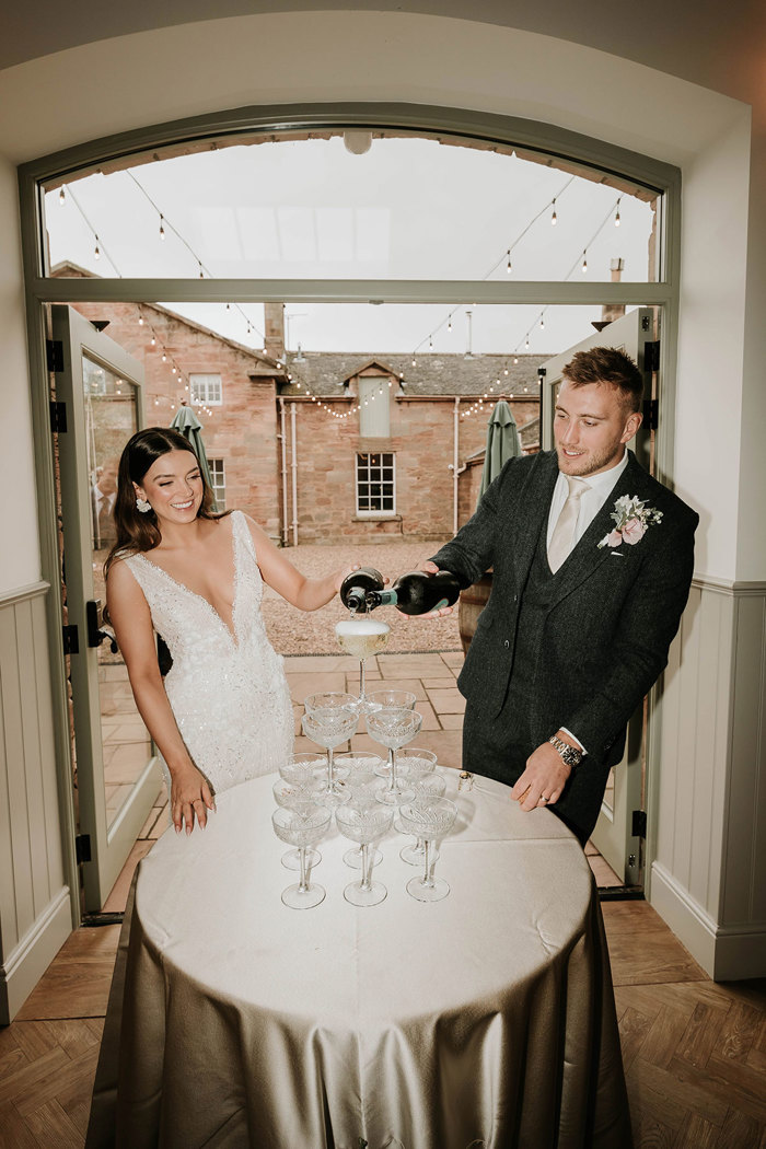 a bride and groom Matt Fagerson pouring champagne into a small tower of coupe glasses in front of a doorway looking onto a courtyard at Dunglass Estate