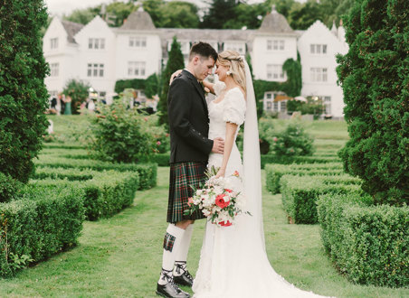 A bride and groom stand outside facing each other in a garden with small hedges and fir trees in front of their venue