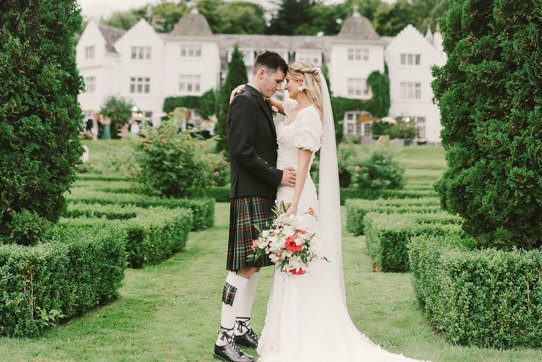 A bride and groom stand outside facing each other in a garden with small hedges and fir trees in front of their venue