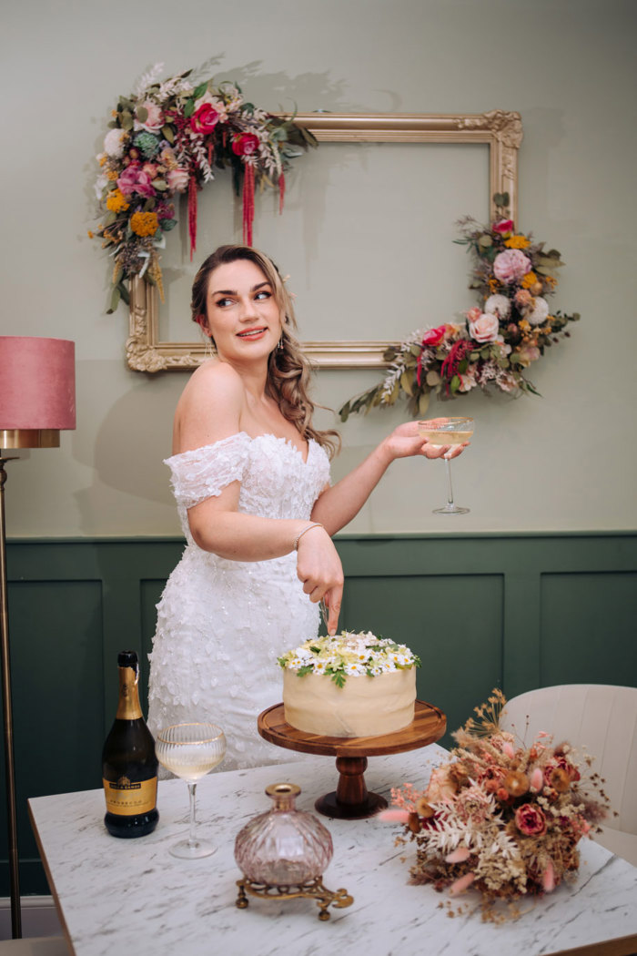 bride in an off the shoulder wedding dress holds a coupe of champagne in one hand and starts to slice a cake with the other