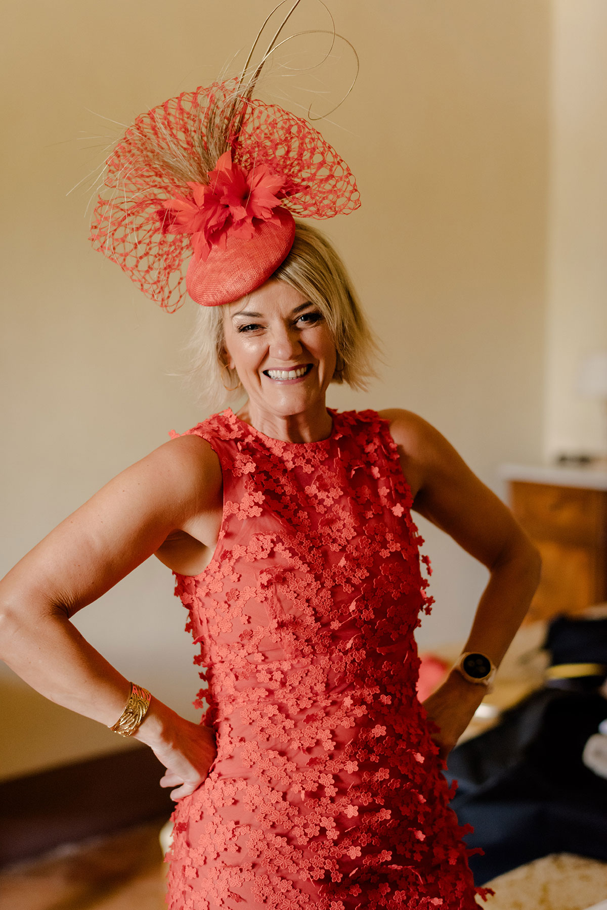 blonde woman with bob smiles and places hands on her hips, wearing a red-orange dress with 3D flowers and a matching percher hat decorated with veiling, quills and feather flowers 