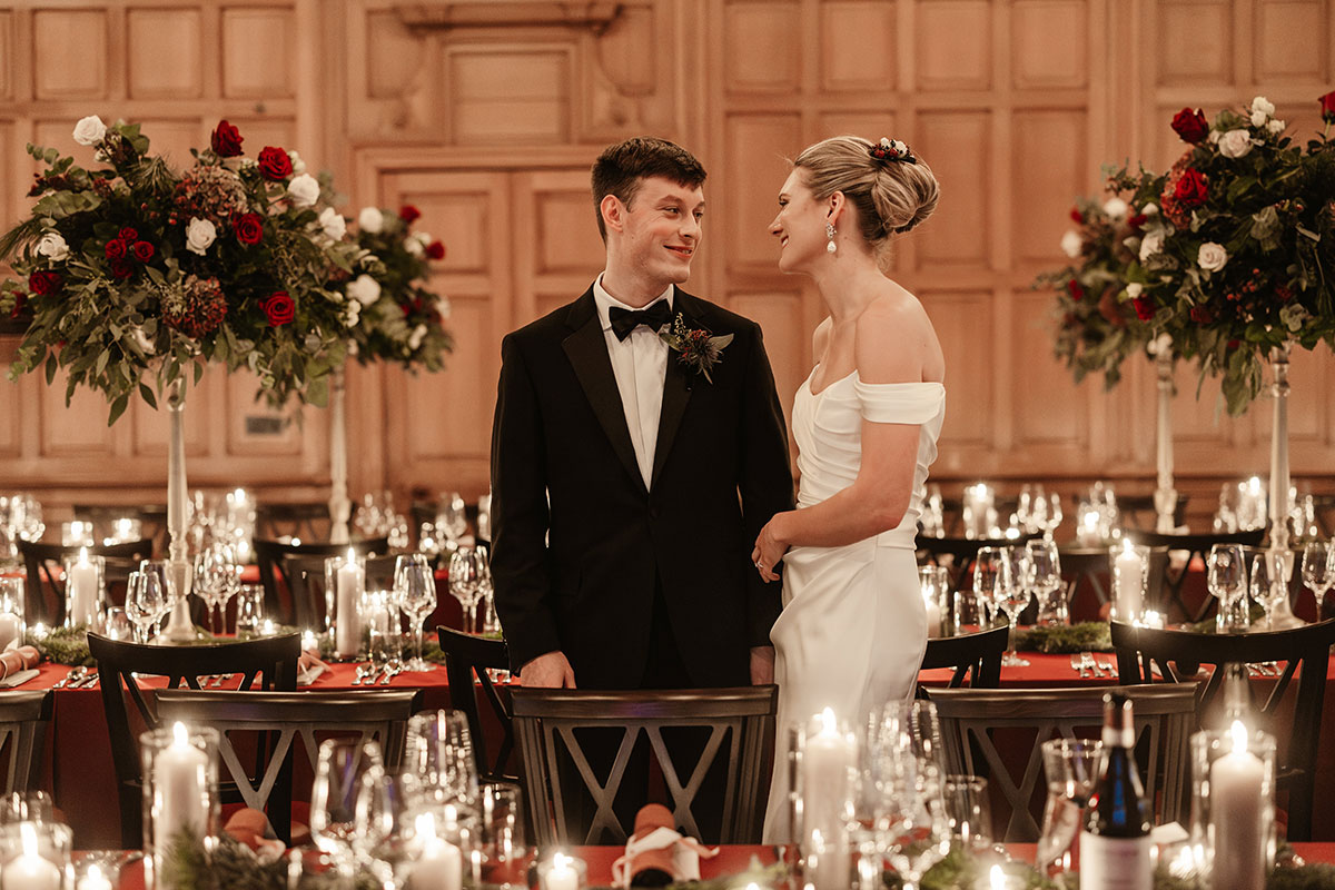 Bride and groom smiling at each other while standing between tall red-and-white floral centrepieces in a candlelit hall.