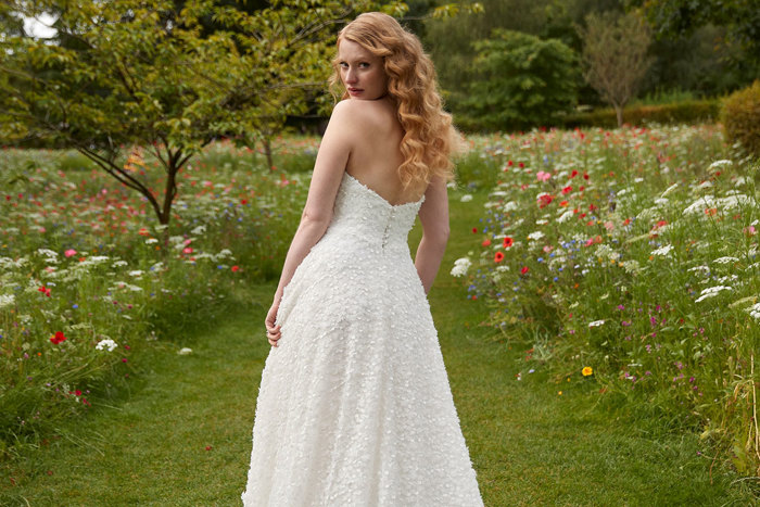 curly haired bride looks back over her shoulder while standing in a grassy flower field wearing a strapless wedding dress