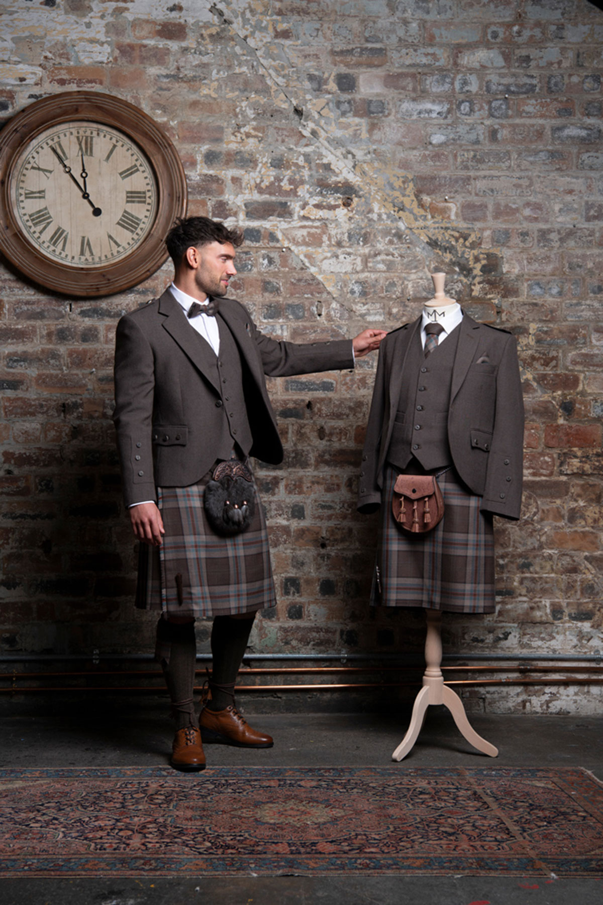 a model wearing a Jura Mist tartan kilt from MacGregor and MacDuff. There is a rustic brick wall and large clock in the background.
