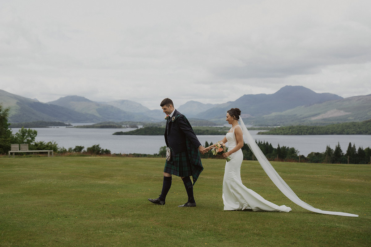 Newly married couple holding hands on scenic lawn overlooking Scottish loch and rolling hills