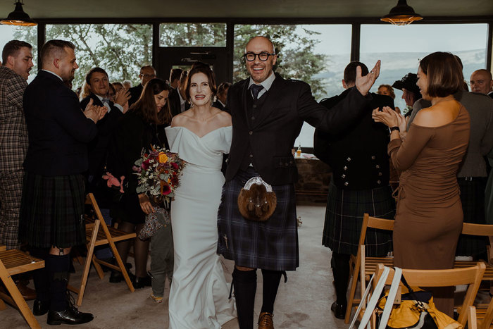 groom in blue, grey and white tartan kilt outfit and bride in off the shoulder white wedding dress holding bouquet under one arm, walk up the aisle between wooden chaired rows of guests stood clapping