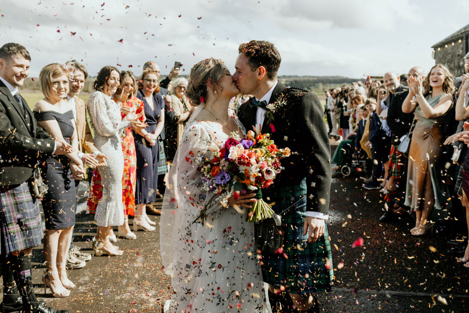 guests smile and throw confetti over bride and groom kissing on their wedding day at bachilton barn