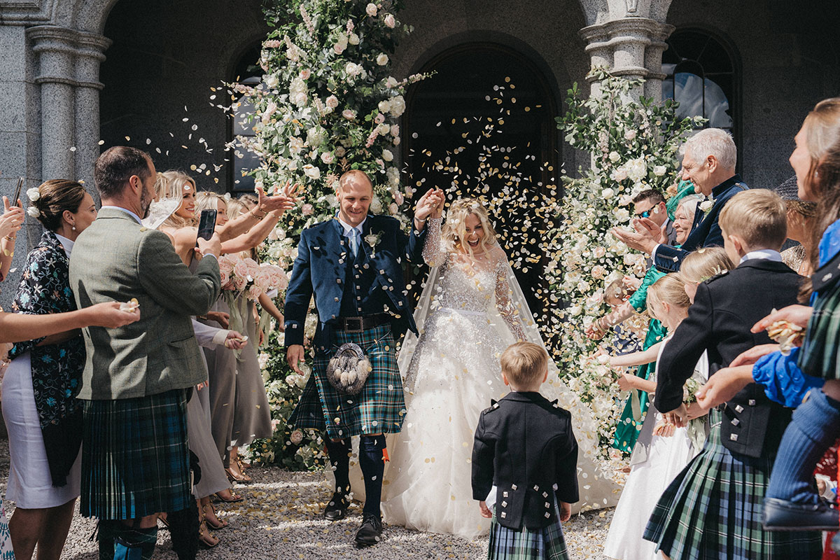 Newlyweds walking through a shower of confetti outside a castle, surrounded by cheering guests