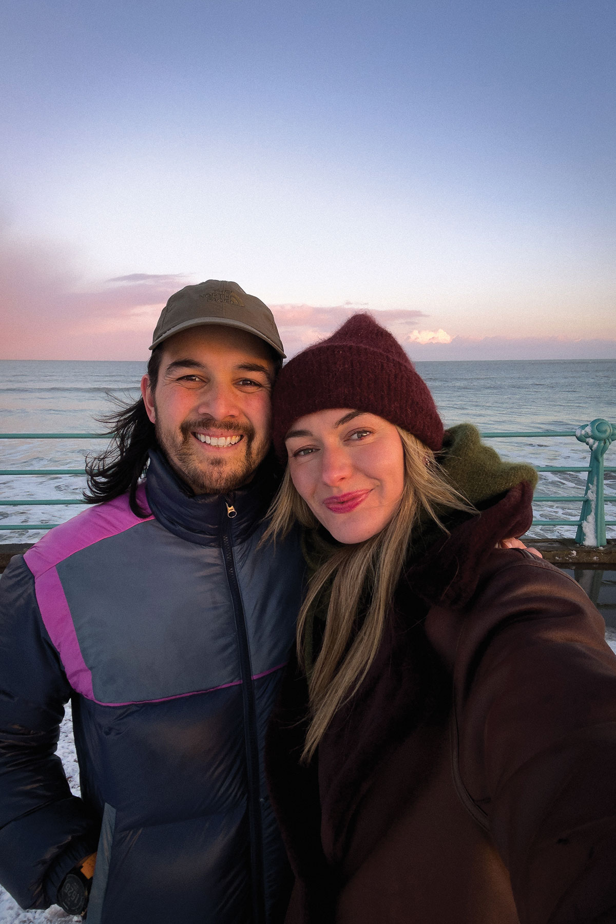 Couple taking a seaside selfie at sunset on a pier; both wear winter hats and jackets with the ocean and pink sky in the background