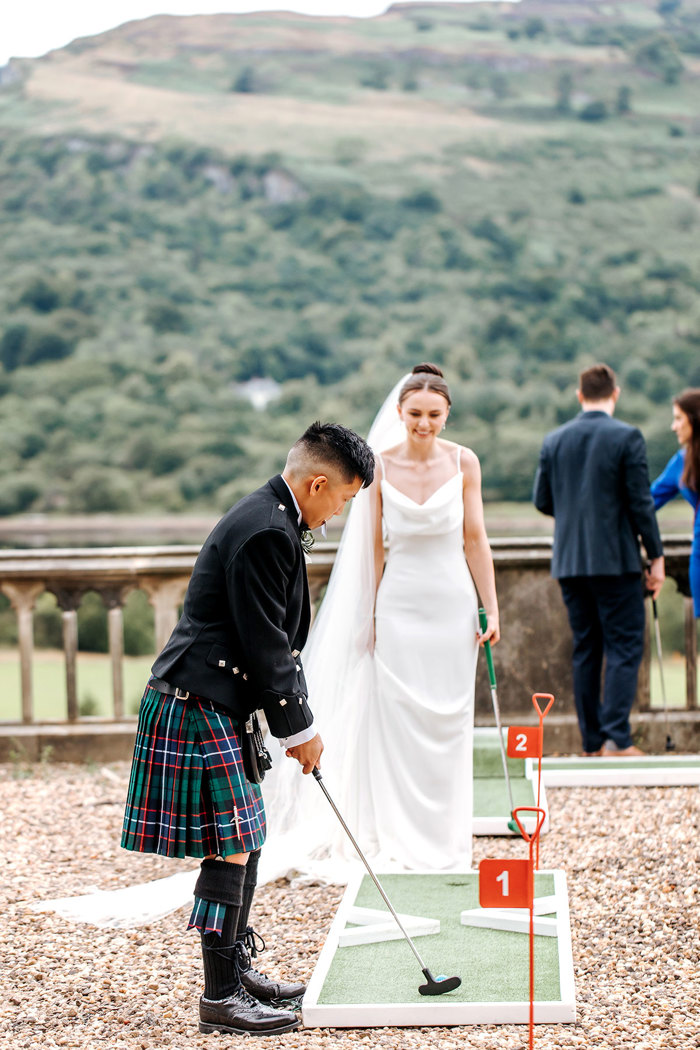 groom wearing a kilt plays mini golf during wedding at mar hall with bride watches
