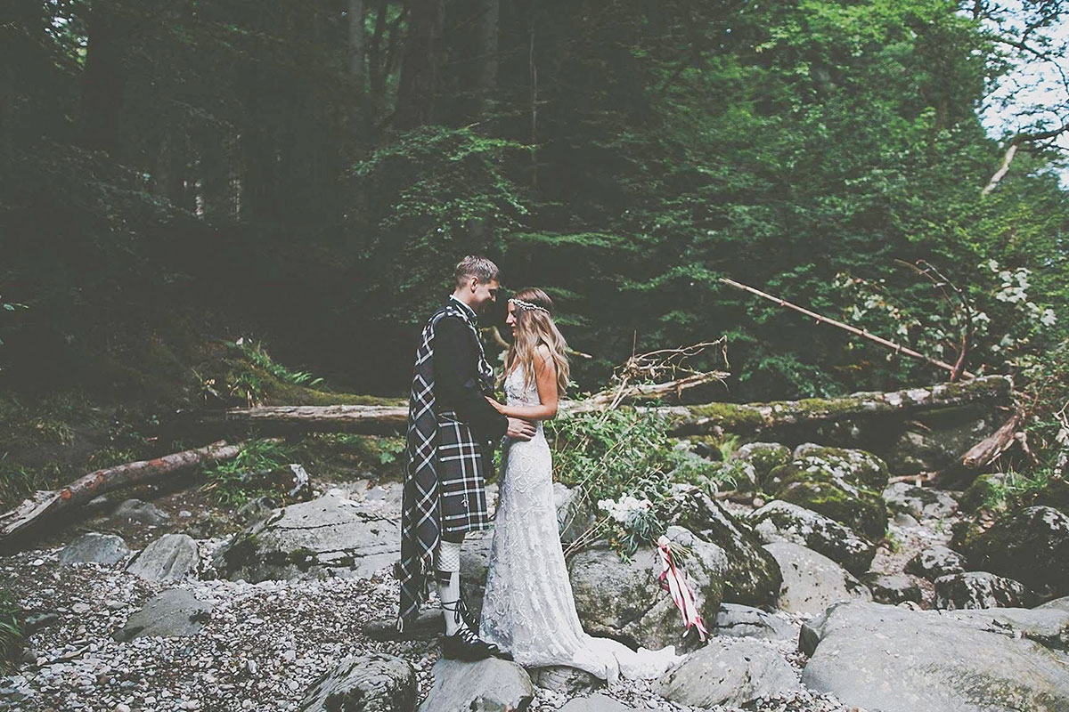 Bride and groom standing together on rocks in a forest setting; the bride wears a fitted lace gown and the groom wears a dark kilt outfit, with a bouquet resting nearby