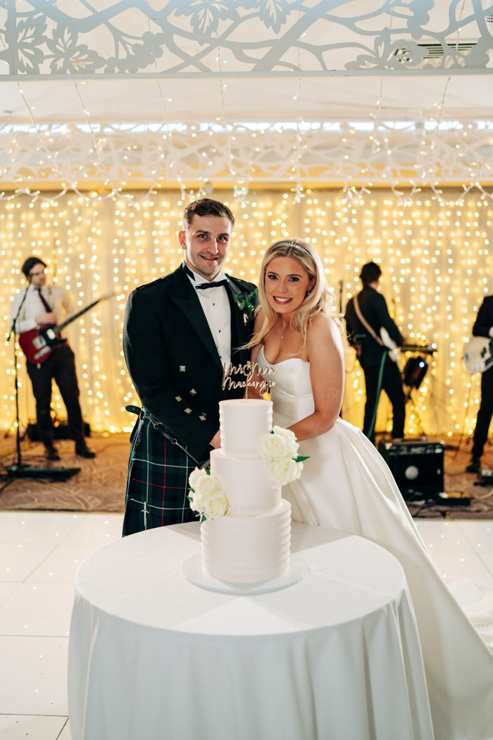 a bride and groom cutting a wedding cake. Behind them, a wedding band play and there's a wall of white sparkly lighting