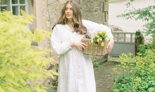 a brunette woman in a long white dress stands outside a stone house holding a basket of yellow tulips