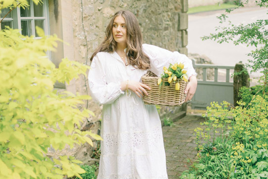 a brunette woman in a long white dress stands outside a stone house holding a basket of yellow tulips