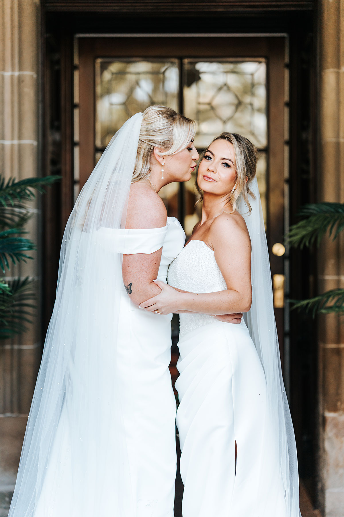 Romantic portrait of one bride kissing her wife's cheek outside the venue as they embrace in matching white gowns and veils