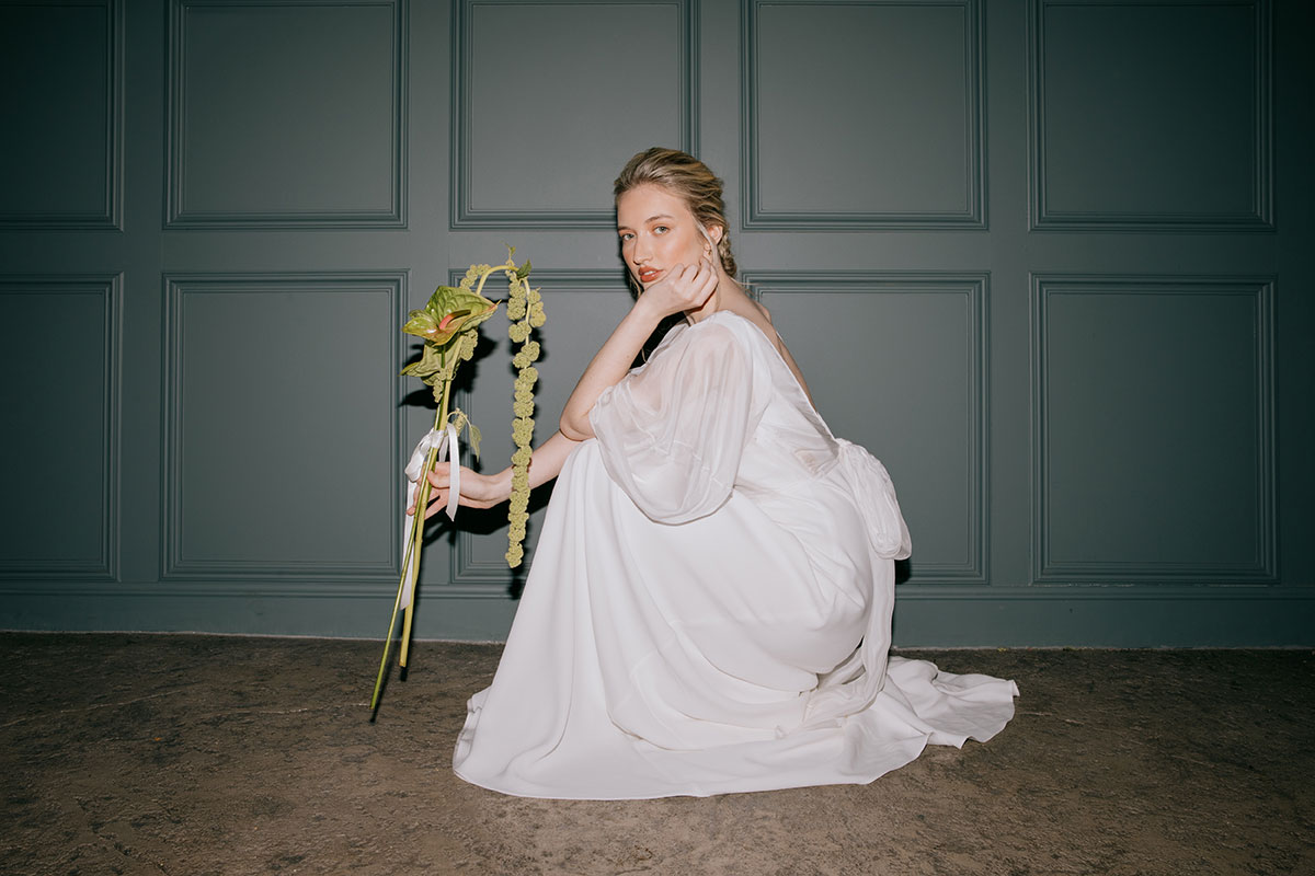 A bride in a flowing white dress crouches in front of a dark teal panelled wall, holding a long-stemmed green anthurium flower.
