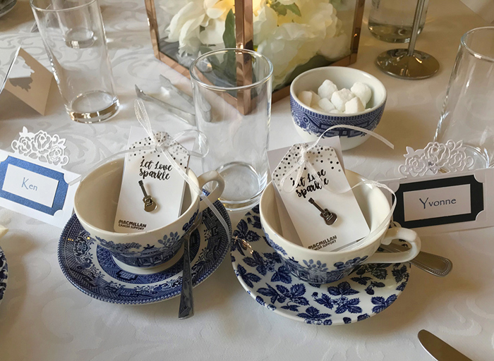 Two tea cups which are white china with a blue pattern sitting on a table next to two placecards and each cup has a silver guitar-shaped pin on a cardboard backing inside it