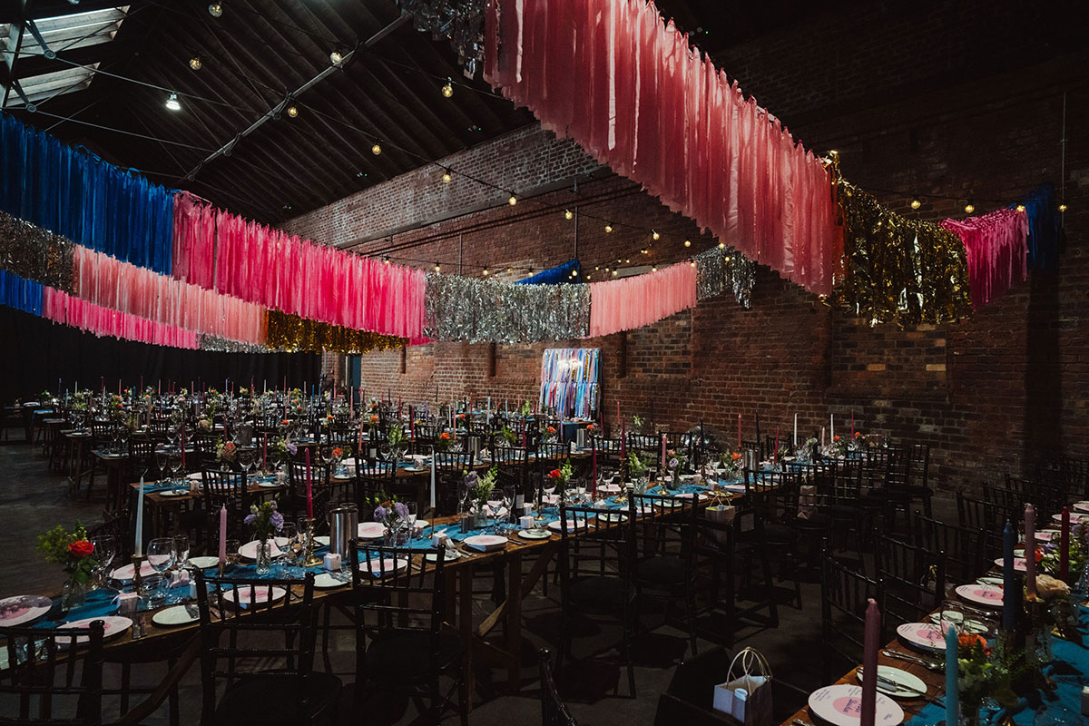 Engine Works wedding reception setup with long banquet tables and dramatic pink, blue and metallic ceiling streamers overhead.