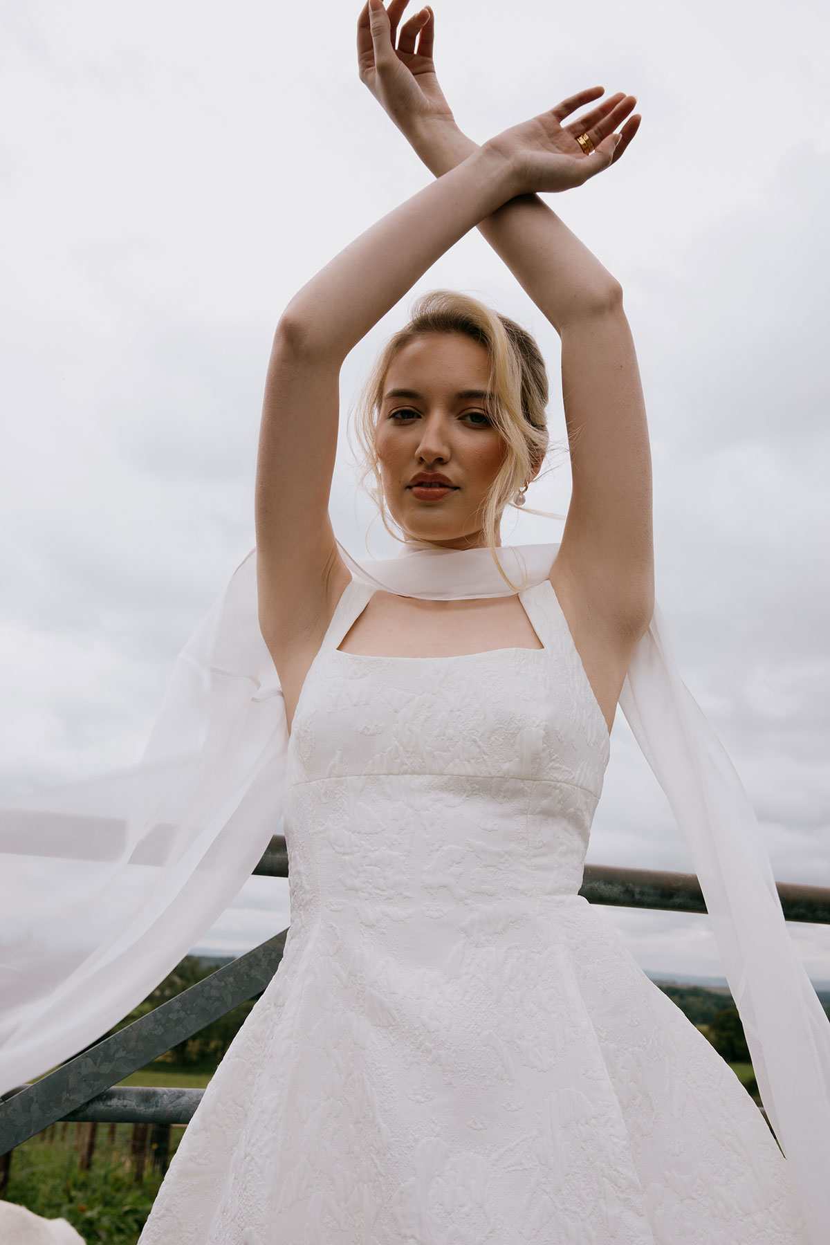 A bride in a square-neck white gown raises her arms while standing at a farm gate, with fields and cloudy skies behind her.