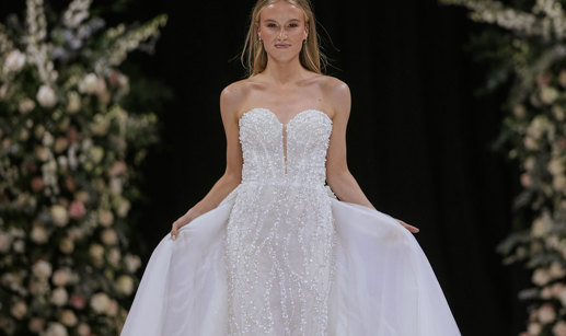 a model wearing a white strapless wedding dress on a catwalk. There are two tall flower displays in the background