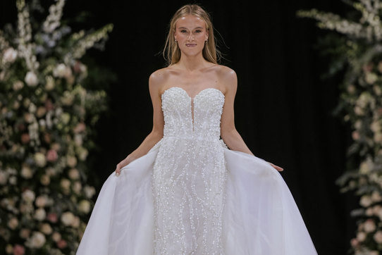 a model wearing a white strapless wedding dress on a catwalk. There are two tall flower displays in the background