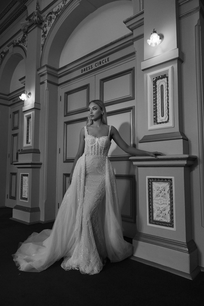a black and white photo of a bride in a wedding dress with a glittery corset bodice and slim fitting skirt with attached overskirt stands outside doors that say 'Dress Circle' above them