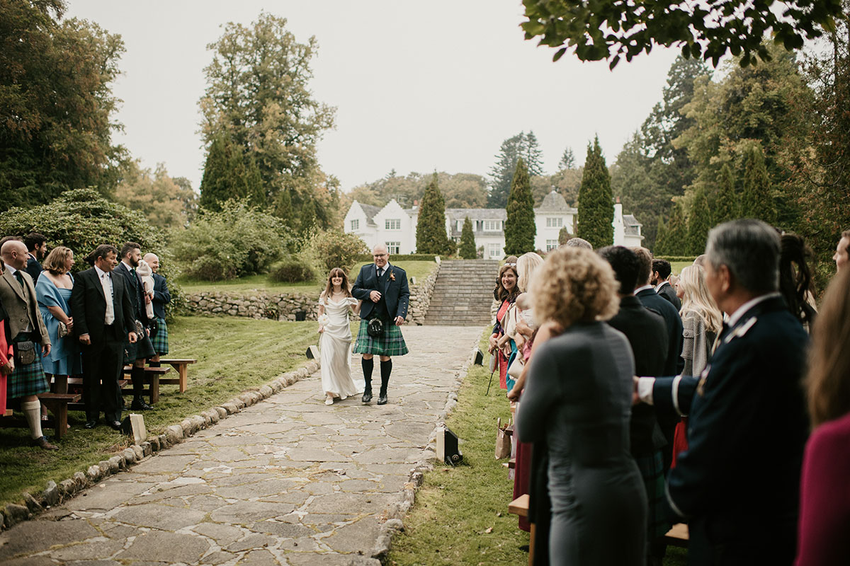 a bride walking down aisle on arm of man in a kilt at outdoor wedding ceremony at Achnagairn Castle