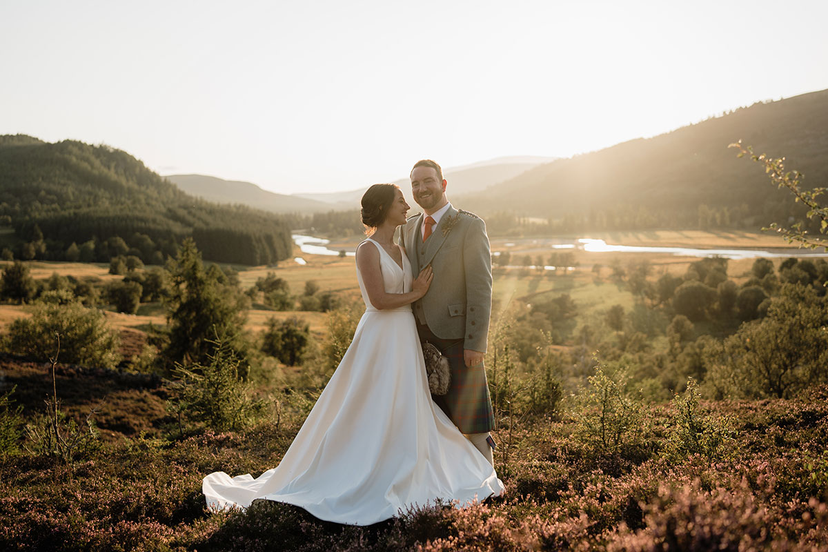 Bride in a flowing white gown and groom in a grey tweed jacket and tartan kilt stand together on a heather-covered hillside, overlooking a winding river and wooded Highland valley in golden evening light