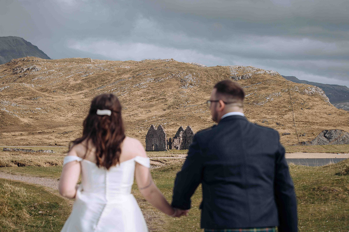 Bride and groom looking towards ruined building below mountain scenery in the Scottish Highlands.