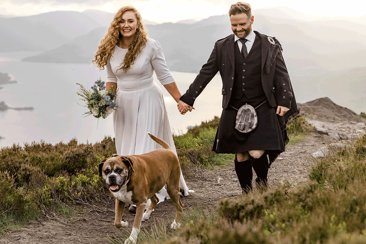 Bride and groom walk hand in hand with their boxer dog along a mountain path after their Scottish elopement ceremony